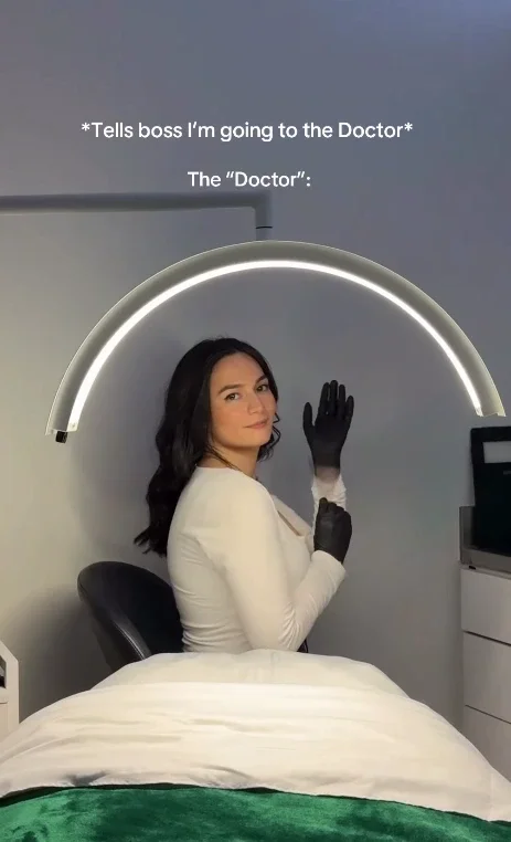 Woman in a white top and black gloves sits under a curved light in a clinic setting, looking back at the camera. Text above references telling a boss she’s going to the “doctor.”. - Jón Alan Salon in Nashville and Franklin, TN