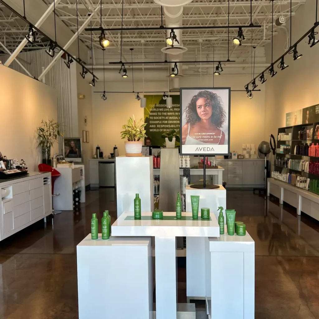 A modern salon interior with product displays, plants, shelves of hair products, and a large poster of a woman advertising Aveda products in the center. - Jón Alan Salon in Nashville and Franklin, TN