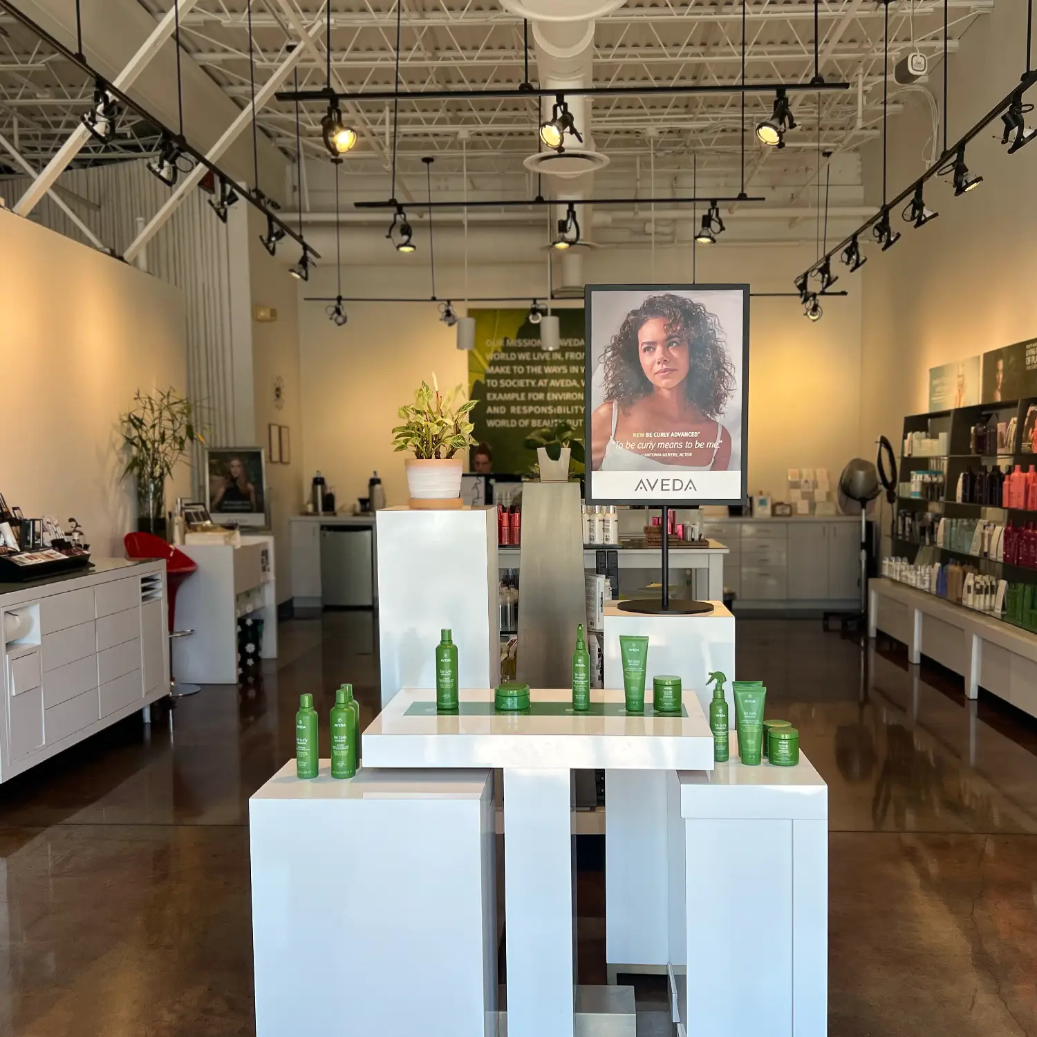 Modern salon interior with white displays showcasing green Aveda hair products, a large poster, plants, and shelves stocked with various beauty products under ceiling track lighting. - Jón Alan Salon in Nashville and Franklin, TN