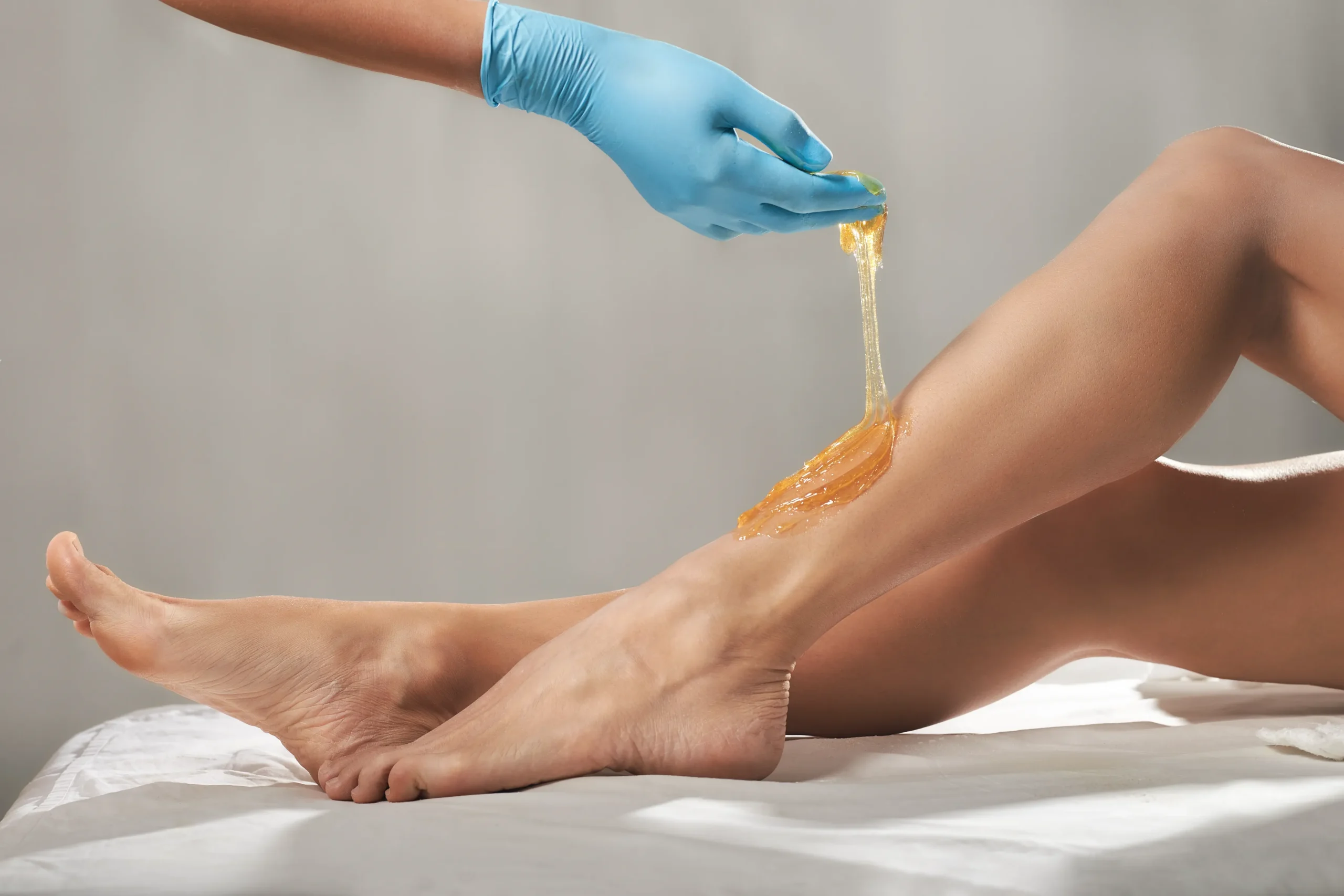 Person wearing blue gloves applying wax to another person’s lower leg for hair removal. Both are on a white surface with a neutral background. - Jón Alan Salon in Nashville and Franklin, TN