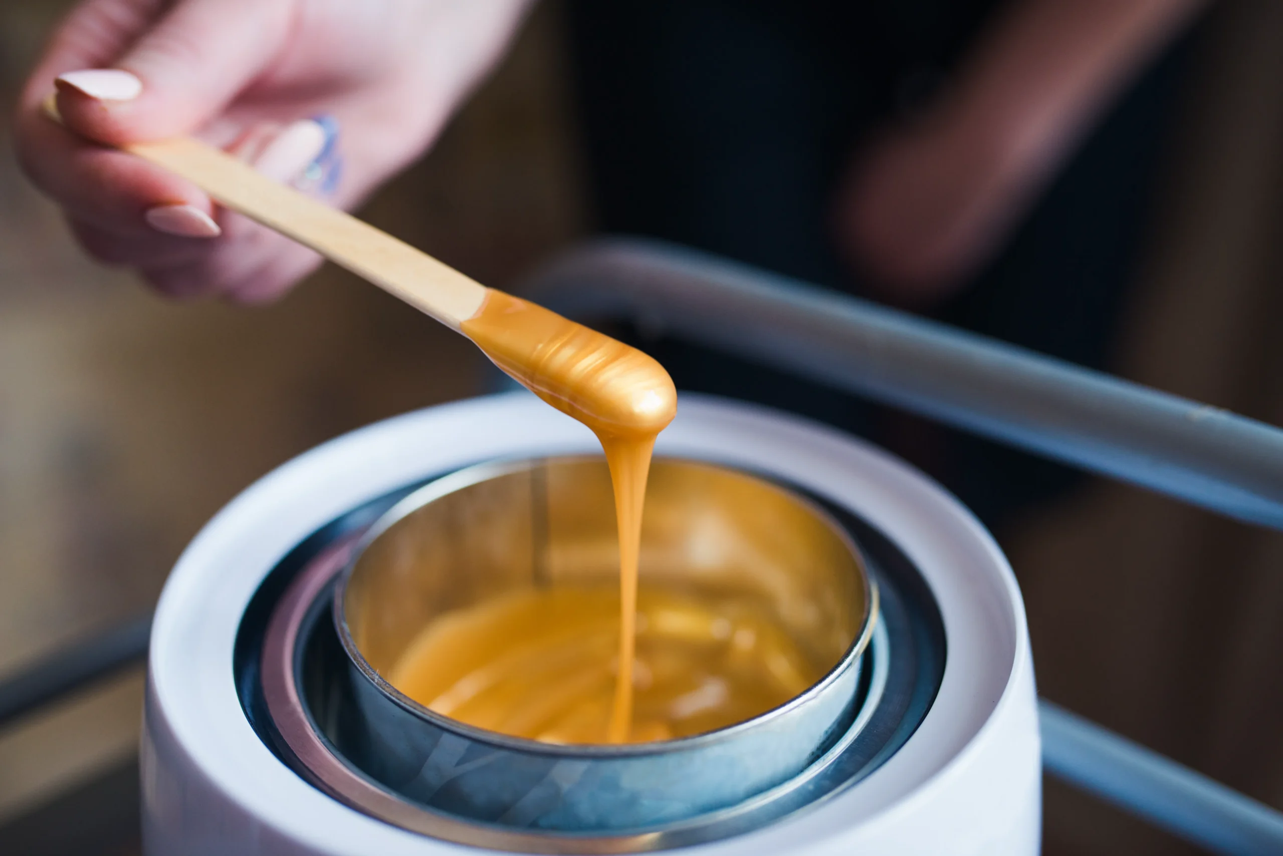 A hand holds a wooden spatula with melted wax dripping from it into a metal container, likely for hair removal. - Jón Alan Salon in Nashville and Franklin, TN